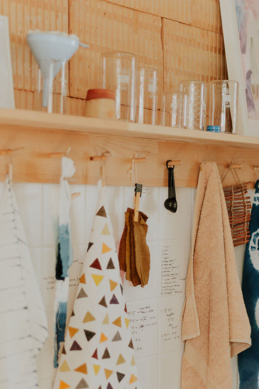 Wooden shelf with various items including a patterned apron, kitchen utensils, and towels in DOT 'N' POT natural dye studio