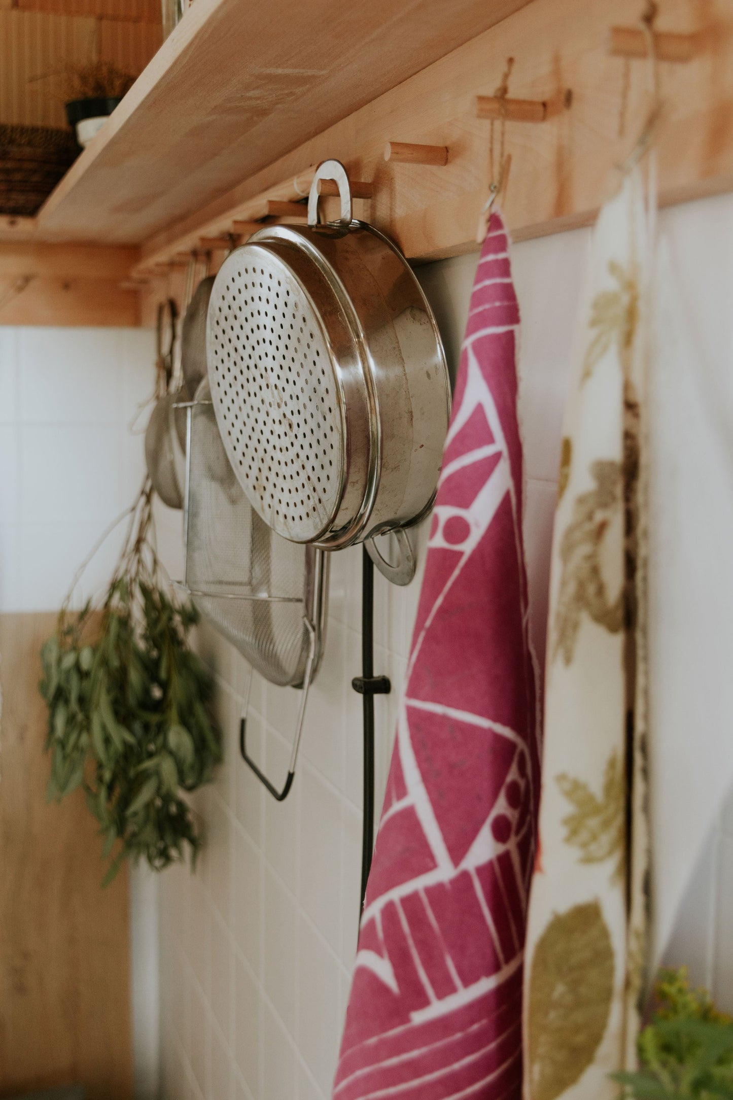 Metal colander hanging on a wooden shelf with kitchen towels and herbs in the background of a natural dye DOT 'N' POT studio